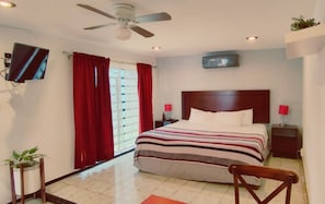 A hotel room with a bed, a ceiling fan, a TV, red curtains, a window, and a small plant. - Suites del Sureste (Mérida)
