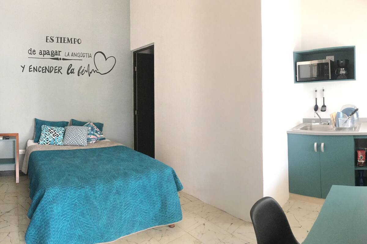 A bedroom with a bed, a bedside table, a wall decoration, a kitchenette with a microwave, and a refrigerator.