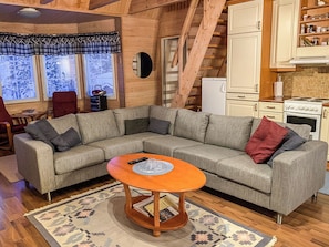 A cozy living room with a sectional sofa, a small wooden coffee table, and a kitchen area with white cabinets and a stove.