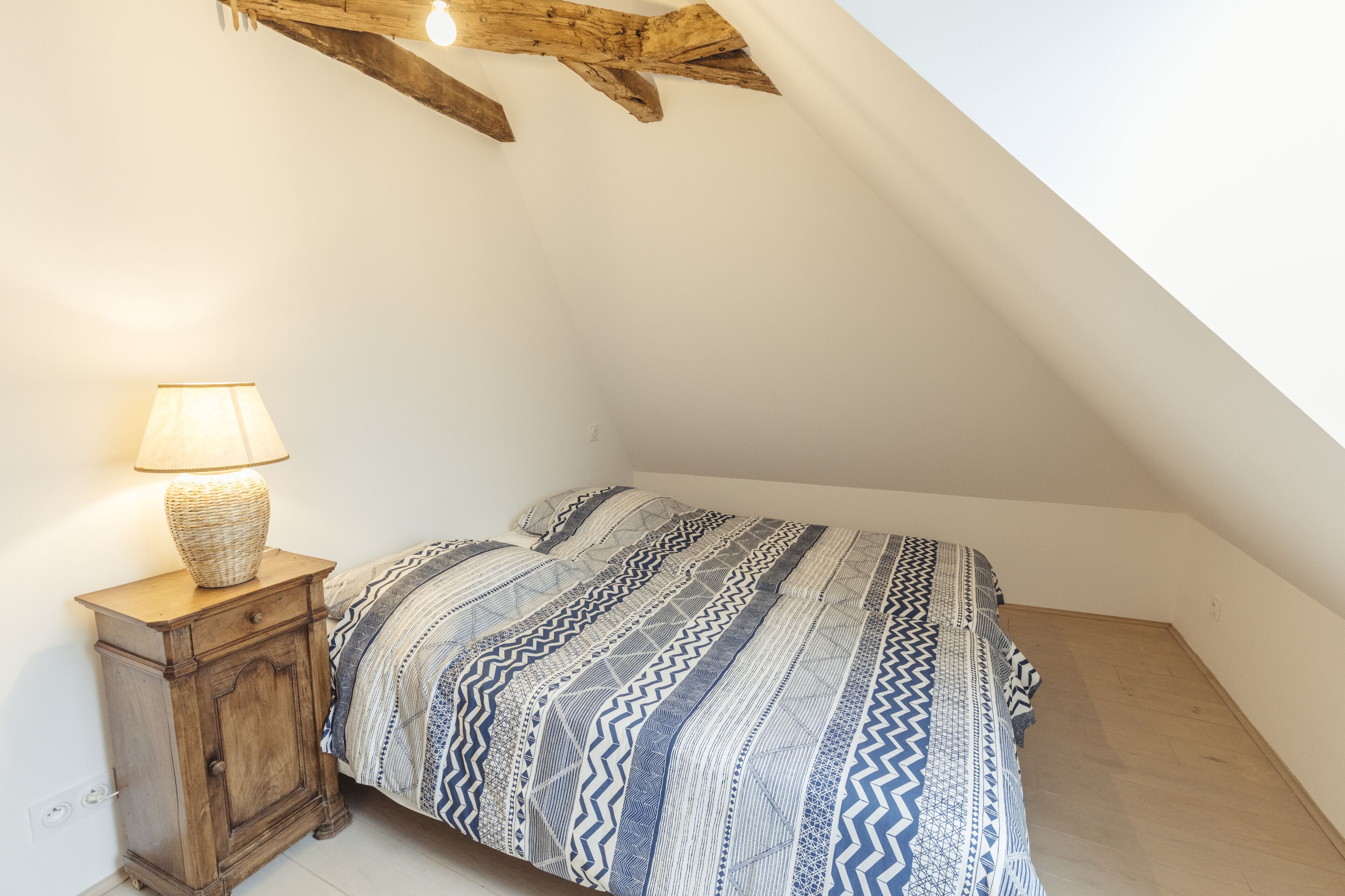 Ein Schlafzimmer mit schrägverdeckter Decke, einem Nachttisch aus Holz mit Lampe sowie einem Bett mit gemustertem Bettbezug.
