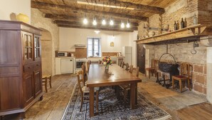 A rustic kitchen with a wooden table, chairs, and a large rug. There is a wooden cabinet, a microwave, and a brick wall with shelves holding bottles. - Gîtes Chez Antonin (Tour-de-Faure)