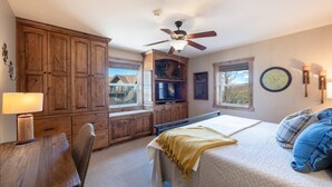 Una camera da letto con un letto, un armadio in legno, una scrivania, una sedia, una ventola a soffitto, una finestra e una sveglia a muro.