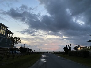 Une rue résidentielle bordée de maisons des deux côtés, un ciel dégagé avec quelques nuages et une étendue d’eau visible au loin.