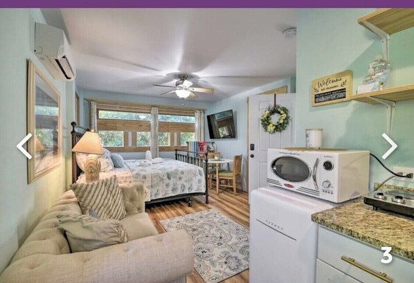 A bedroom with a bed, a bedside table, a lamp, and a window. There is a kitchenette with a microwave and a refrigerator.