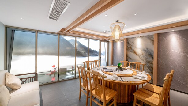 A modern dining area with a round wooden table, wooden chairs, and a large window offering a scenic view of a body of water and mountains. - Bo Yi Hotel (Guangzhou)