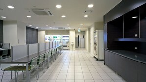 Una cafetería moderna y bien iluminada con mesas y sillas largas, máquinas expendedoras y un refrigerador.