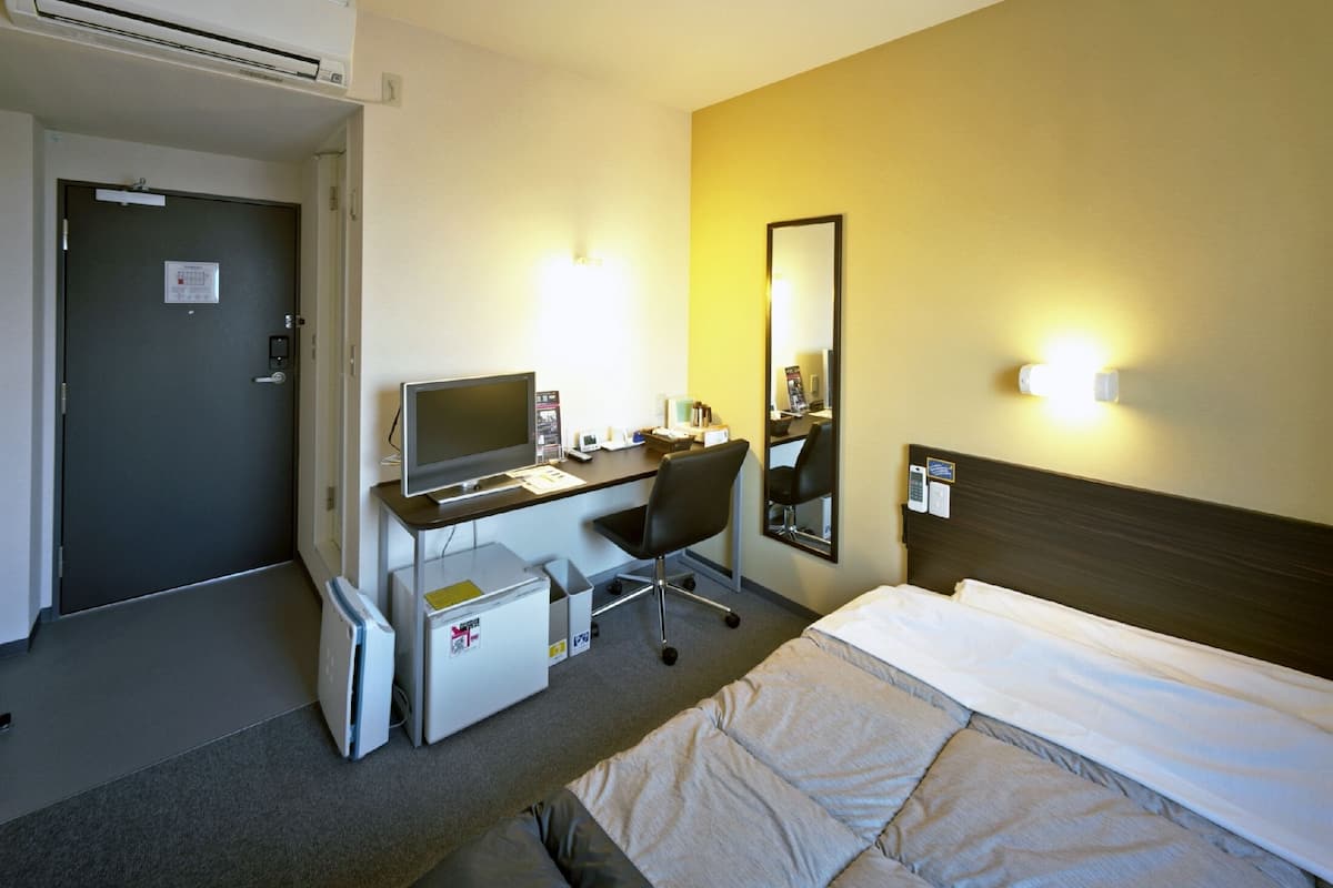 A hotel room with a bed, a desk with a computer, a chair, a small refrigerator, and a mirror on the wall.