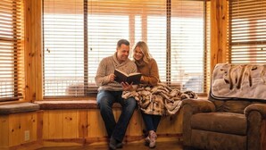 A couple sitting by a window with wooden blinds, a cushioned bench, and a fur throw on a brown sofa. - Cozy and lakefront cabin on Rainbow lake, large fenced deck and 2 grills!!! (Pinetop-Lakeside)