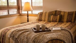 Een bed met een geborduurde spreideken, een dienblad met twee mokken, een boek en een lamp op een nachtkastje.