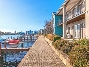 A waterfront property with a paved walkway, a dock with a boat, and modern buildings with balconies. - Waters Edge Apartment with Jetty (Halls Head)