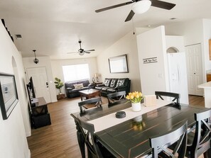 Un vaste espace de vie comprenant une table à manger en bois foncé, des chaises noires, un canapé en cuir noir et un ventilateur de plafond.