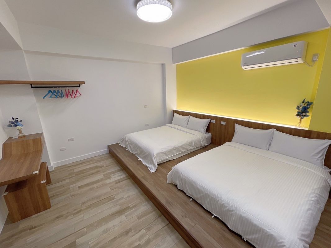 A hotel room with two beds, a wooden desk, a hanging clothes rack, and a yellow accent wall.