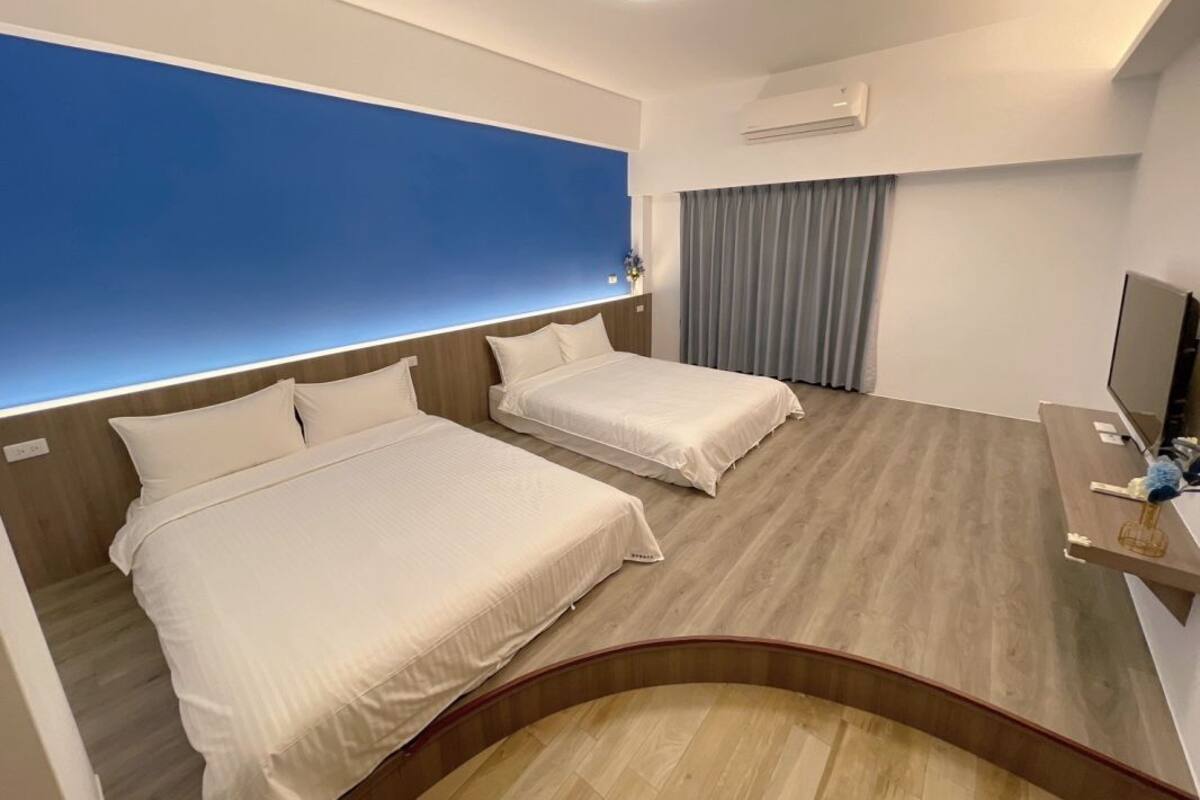 A hotel room with two beds, a desk with a computer, a TV, and a large window with a blue wall.