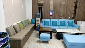 Una sala moderna con un sofá color beige, un sofá seccional azul y una mesa de centro. Se observa un dispensador de agua y un televisor al fondo.