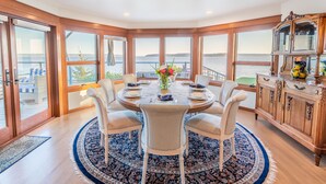 Un comedor con una mesa redonda, seis sillas, un tapete azul y blanco con diseño y vistas al agua a través de amplias ventanas.