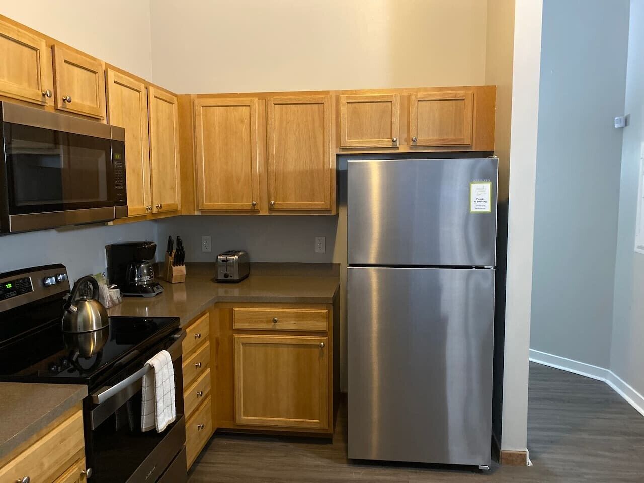 A compact kitchen with wooden cabinets, a stainless steel refrigerator, a microwave, a stove, and a toaster.