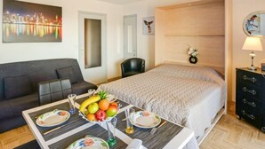 Et hotelværelse med seng, sofa, spisebord med frugt og bestik, et spejl og et bybillede.