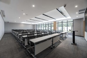 Una sala conferenze con file di tavoli e sedie, ampie finestre e un podio.