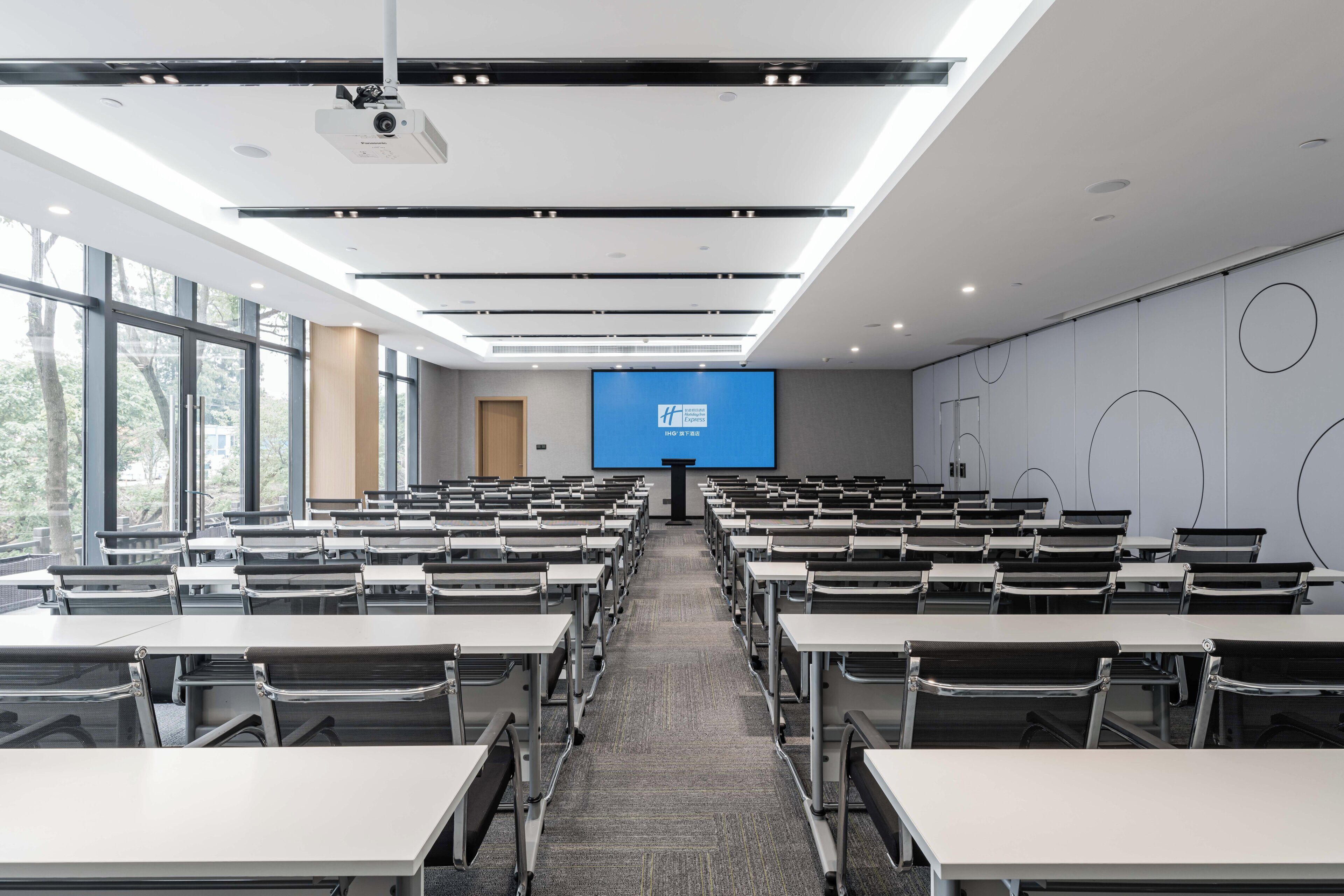 Un salón de conferencias con filas de mesas y sillas, una pantalla grande al frente y un proyector montado en el techo.