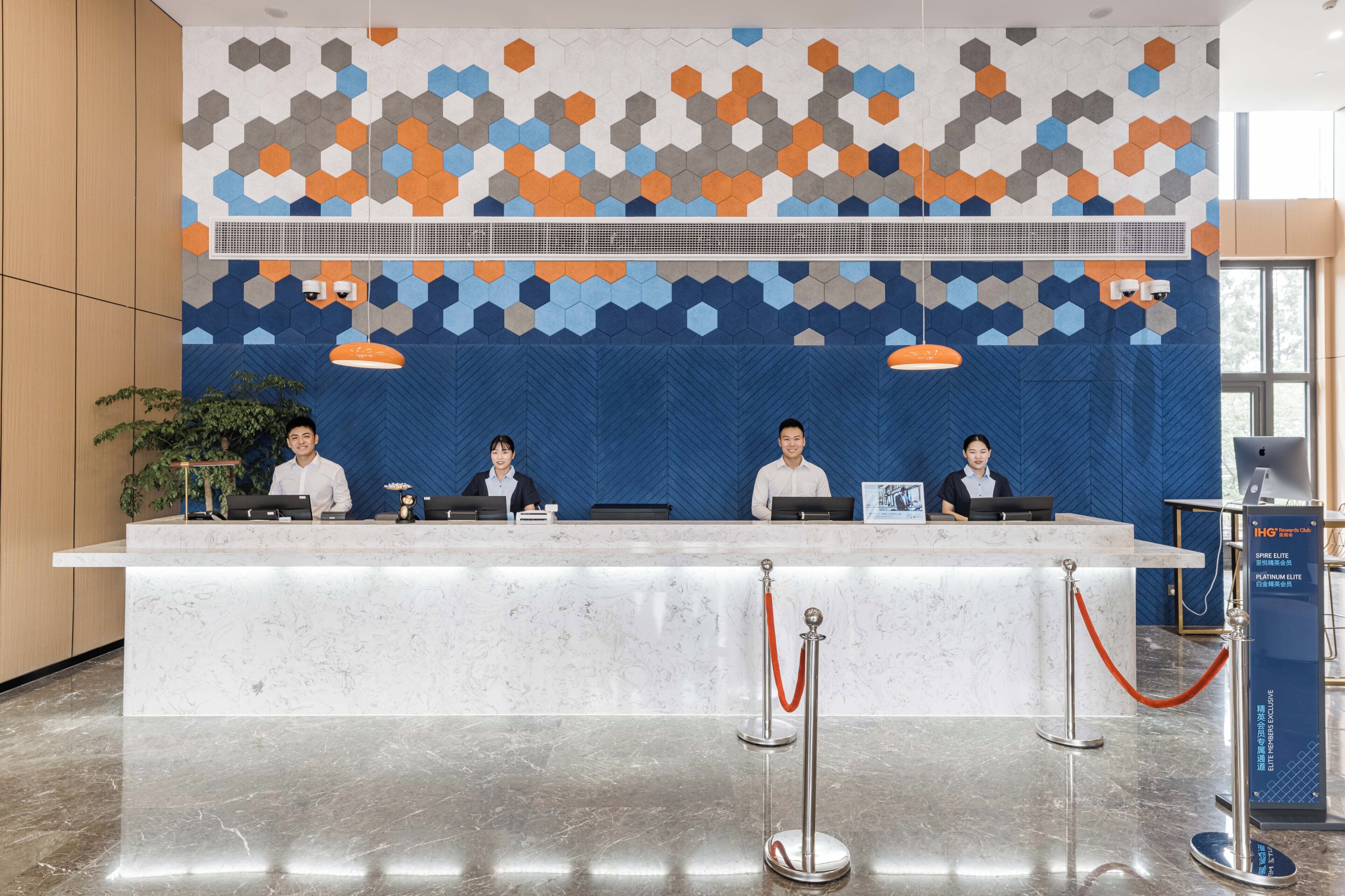 Balcão de recepção de hotel com quatro funcionários, parede estampada em azul e piso de mármore.