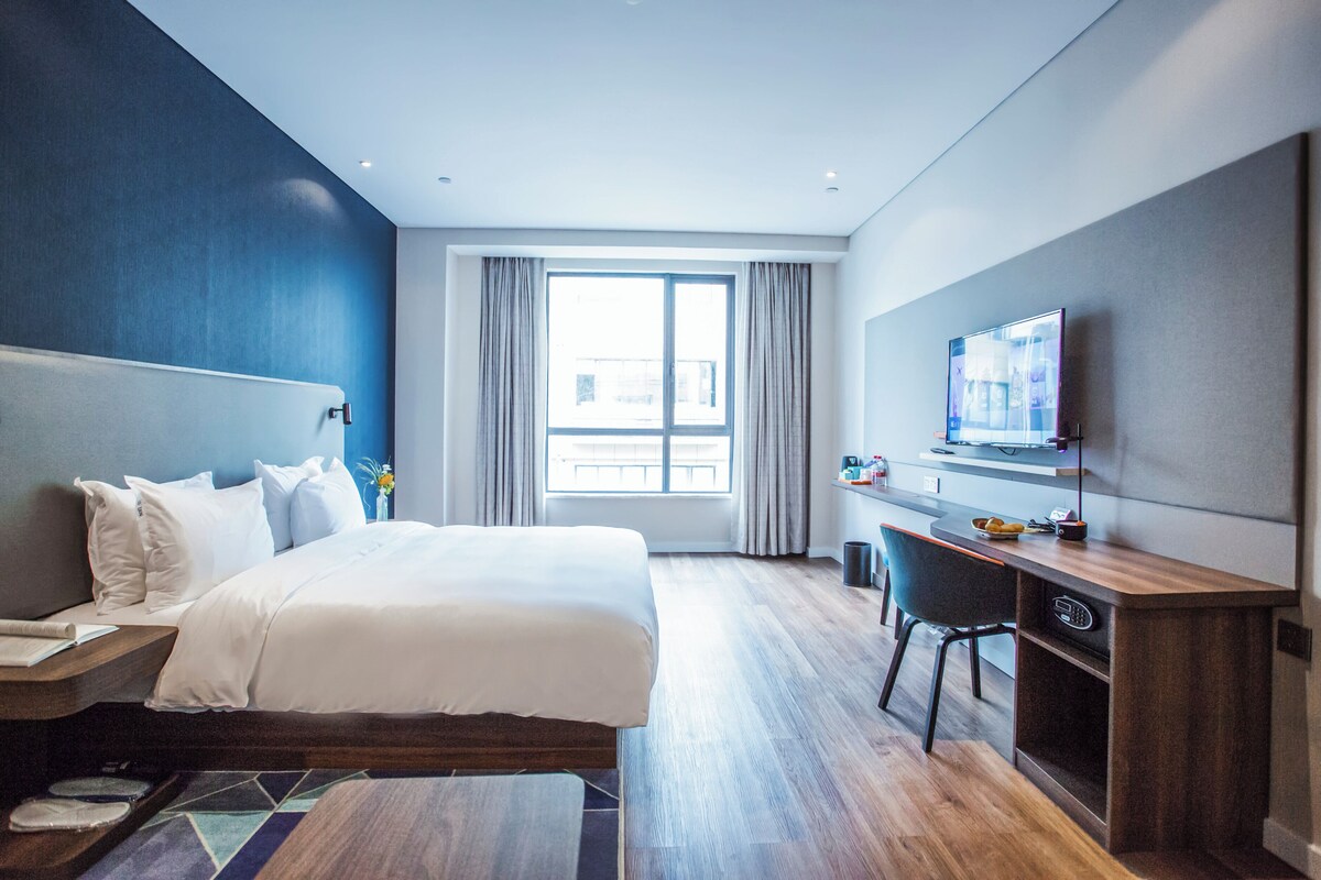 Ein modernes Hotelzimmer mit einem großen Bett, einem Schreibtisch, einem Fernseher und einem Fenster mit Vorhängen.