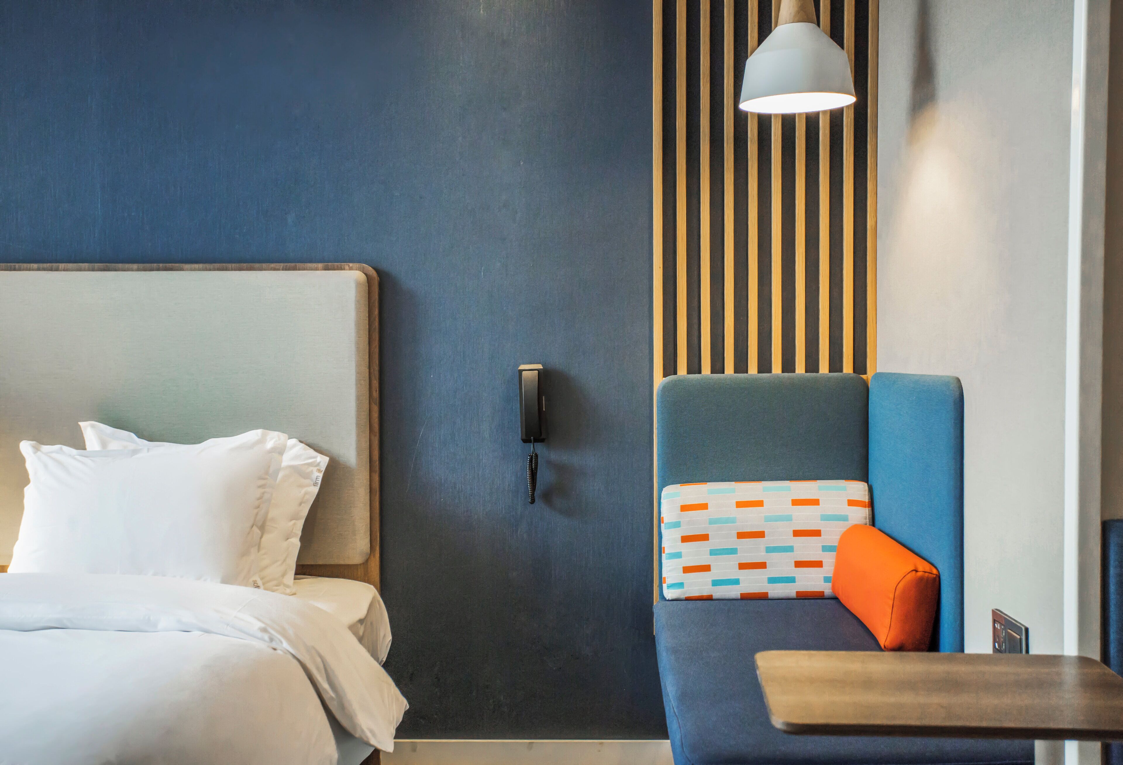 Una habitación de hotel con una cama, una zona de estar con un cojín estampado, una lámpara fijada a la pared y un panel de madera.