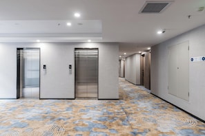 A hotel corridor with three elevators, a patterned carpet, and a sign indicating room numbers.