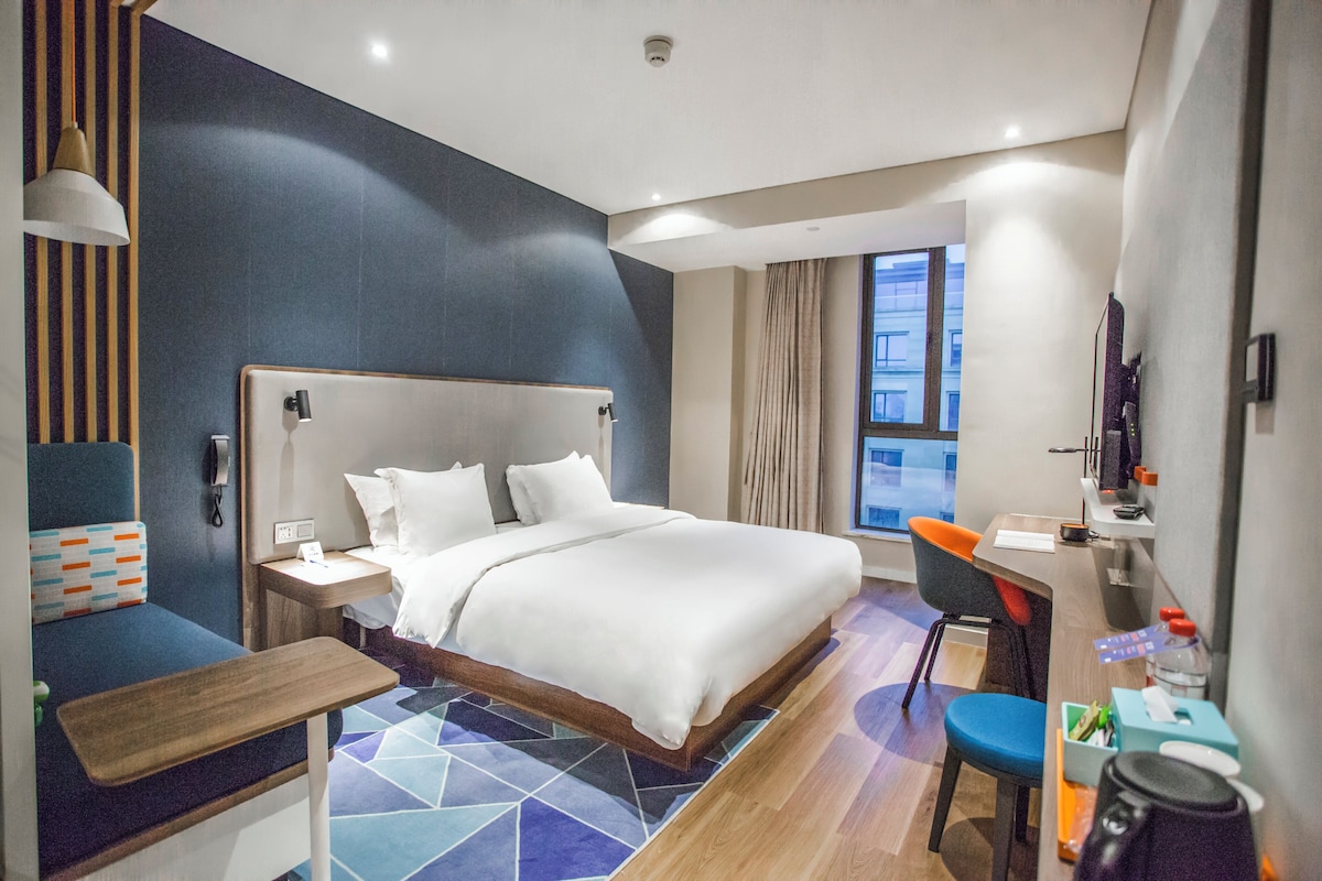 Ein modernes Hotelzimmer mit einem großen Bett, einem Schreibtisch, einem Sessel und Blick auf die Stadt.