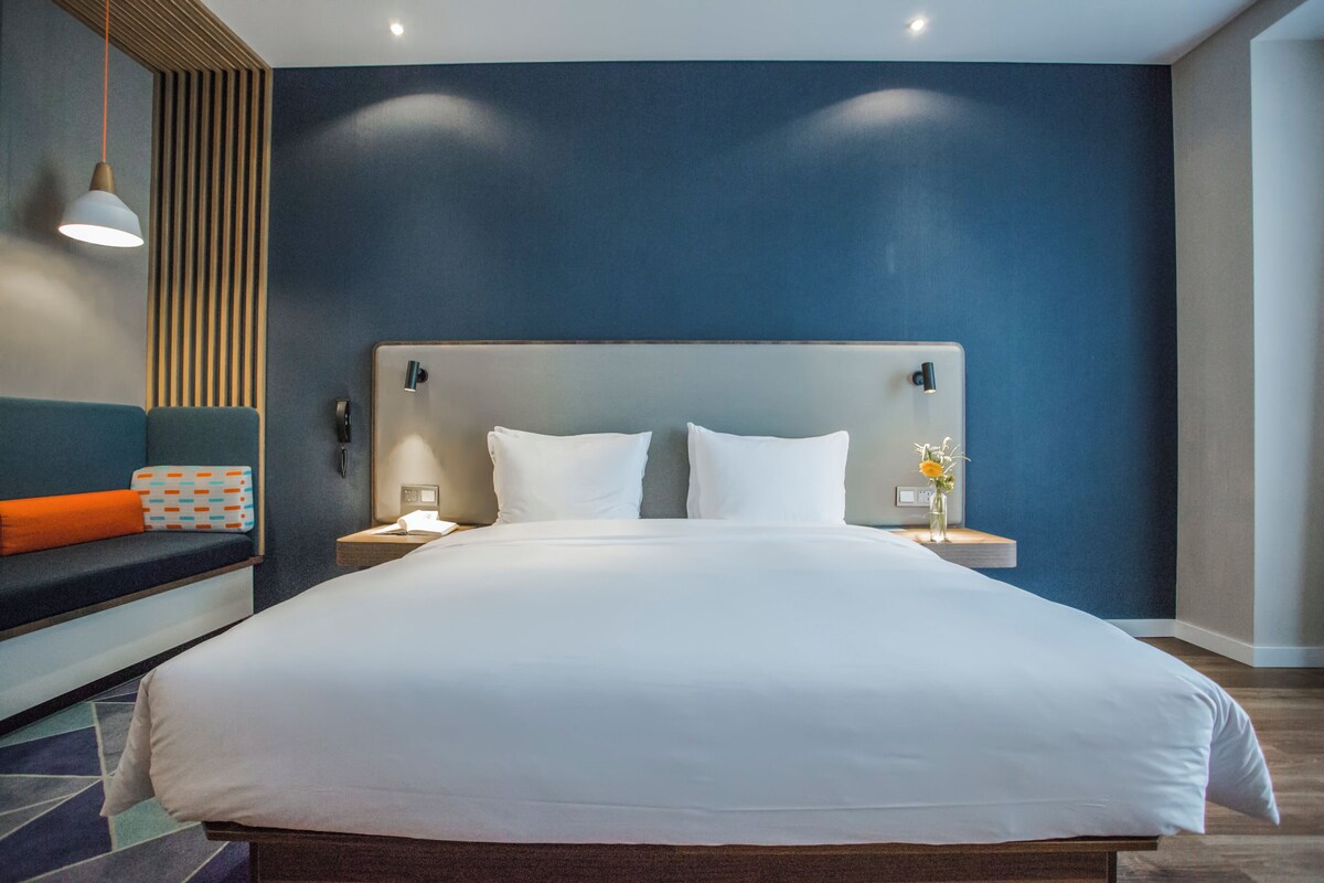 Ein modernes Hotelzimmer mit einem großen Bett, Nachttischen, einer Ablage, einer Wandlampe und einer Sitzecke mit Kissen.