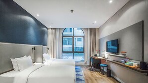 Ein Hotelzimmer mit einem großen Bett, einem Schreibtisch mit Stuhl, einem Fernseher und Blick auf die Stadt durch ein großes Fenster.