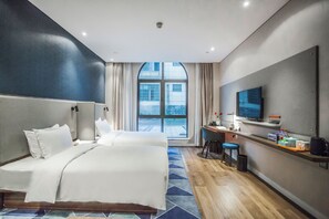 Quarto de hotel com uma cama grande, uma escrivaninha com cadeira, televisão e vista da cidade por uma janela ampla.