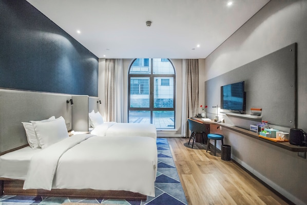 Quarto de hotel com uma cama grande, uma escrivaninha com cadeira, televisão e vista da cidade por uma janela ampla.