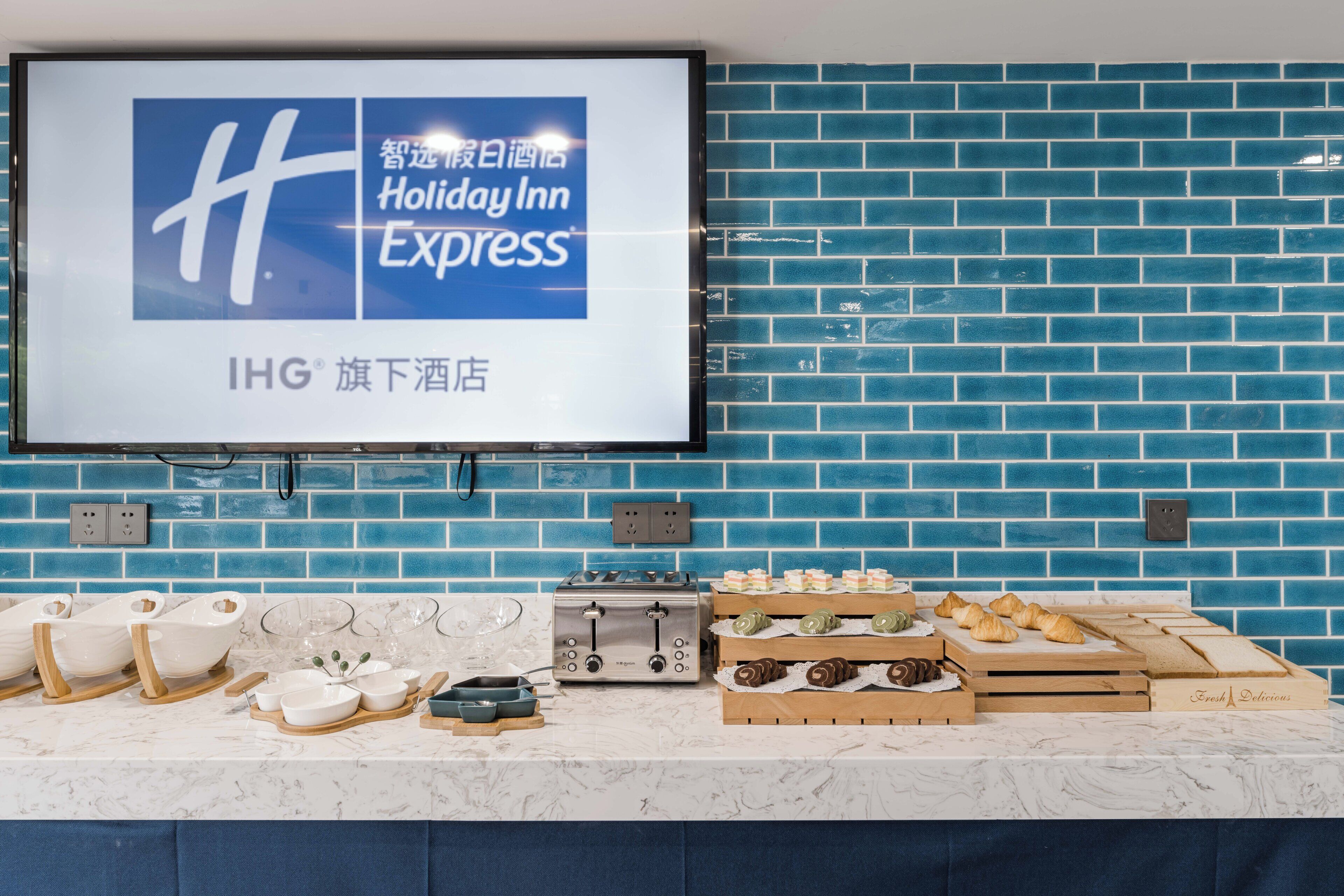 A hotel room with a blue brick wall, a flat-screen TV displaying the Holiday Inn Express logo, a marble countertop with various breakfast items, and electrical outlets.
