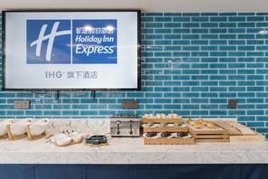 Habitación de hotel con pared de ladrillo azul, un televisor de pantalla plana mostrando el logotipo de Holiday Inn Express, encimera de mármol con diversos productos para el desayuno y enchufes eléctricos.