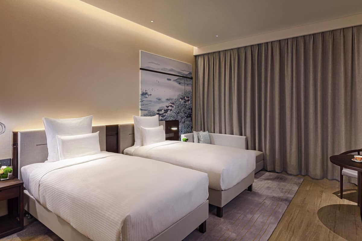A hotel room with two beds, a painting on the wall, a curtain, a small table, and a nightstand with a plant.