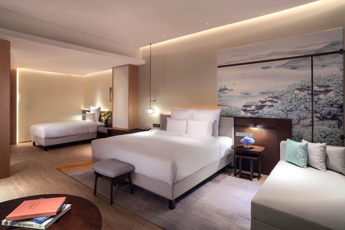 A modern hotel room with a large bed, a smaller sofa, a desk, and a wall-mounted TV.