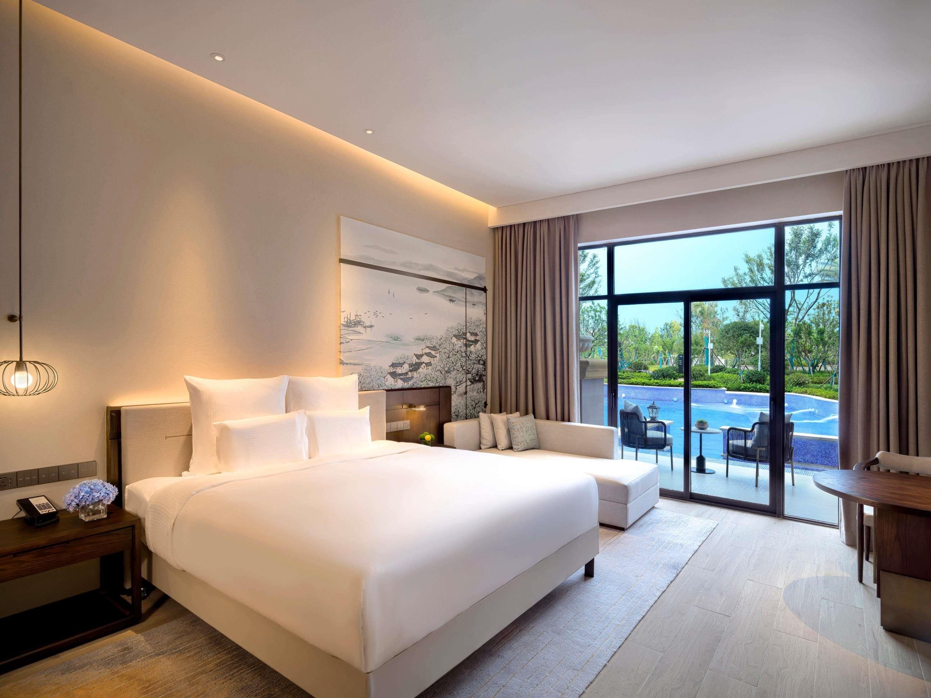 Habitación de hotel moderna con una cama grande, un sofá, una mesa de comedor y vista a una piscina.
