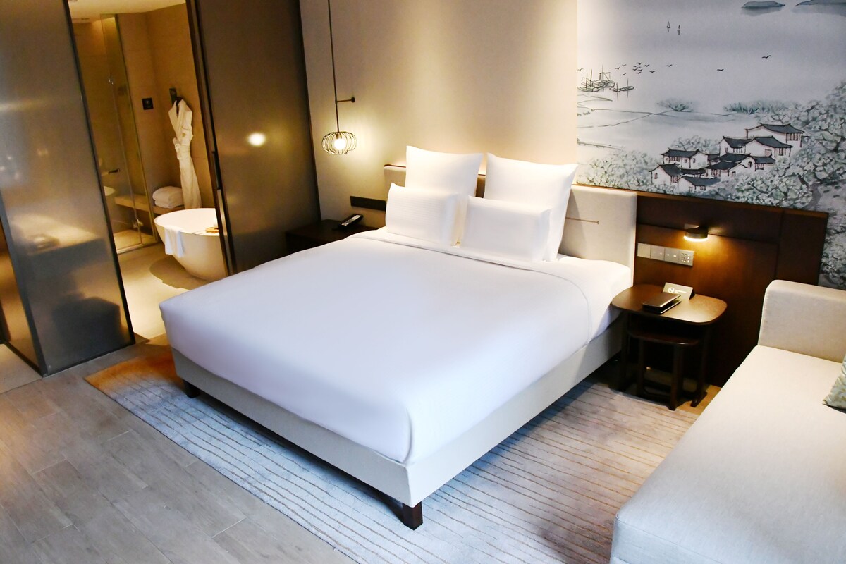 A modern hotel room with a large bed, a bedside table with a lamp, a small table with a phone, and a wall-mounted shelf.