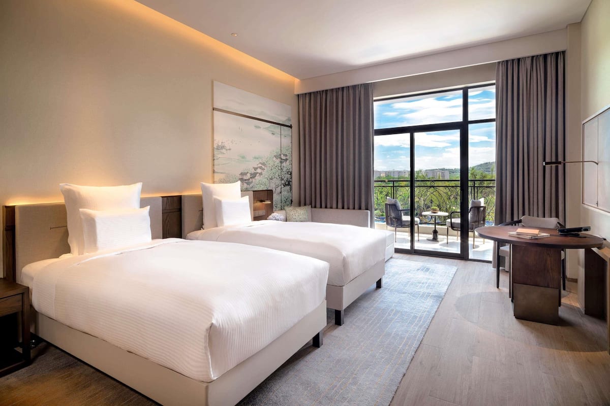A modern hotel room with two beds, a balcony with a view, and a desk with a chair.