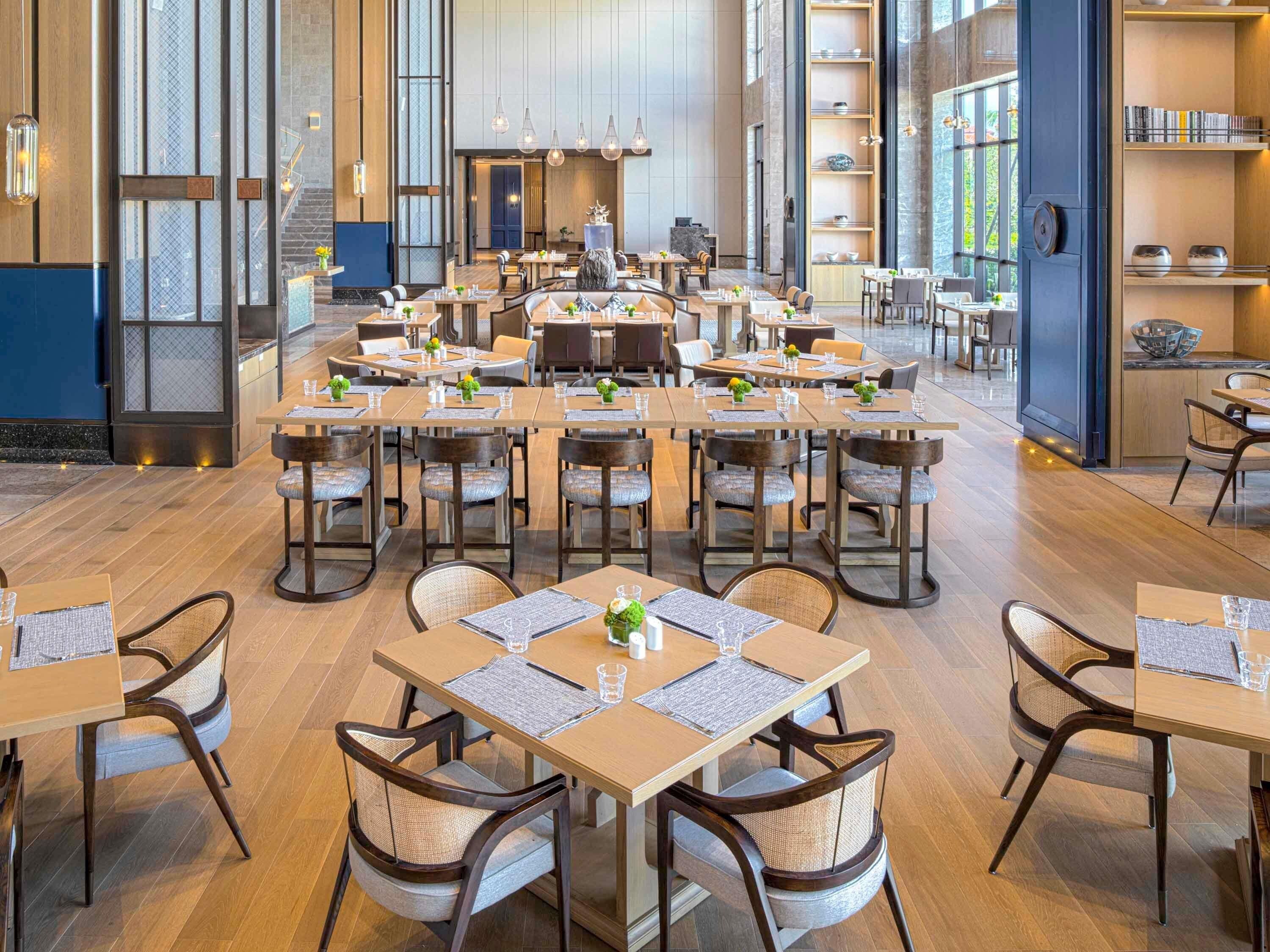 A spacious dining area with multiple tables and chairs, large windows, and a modern interior design.