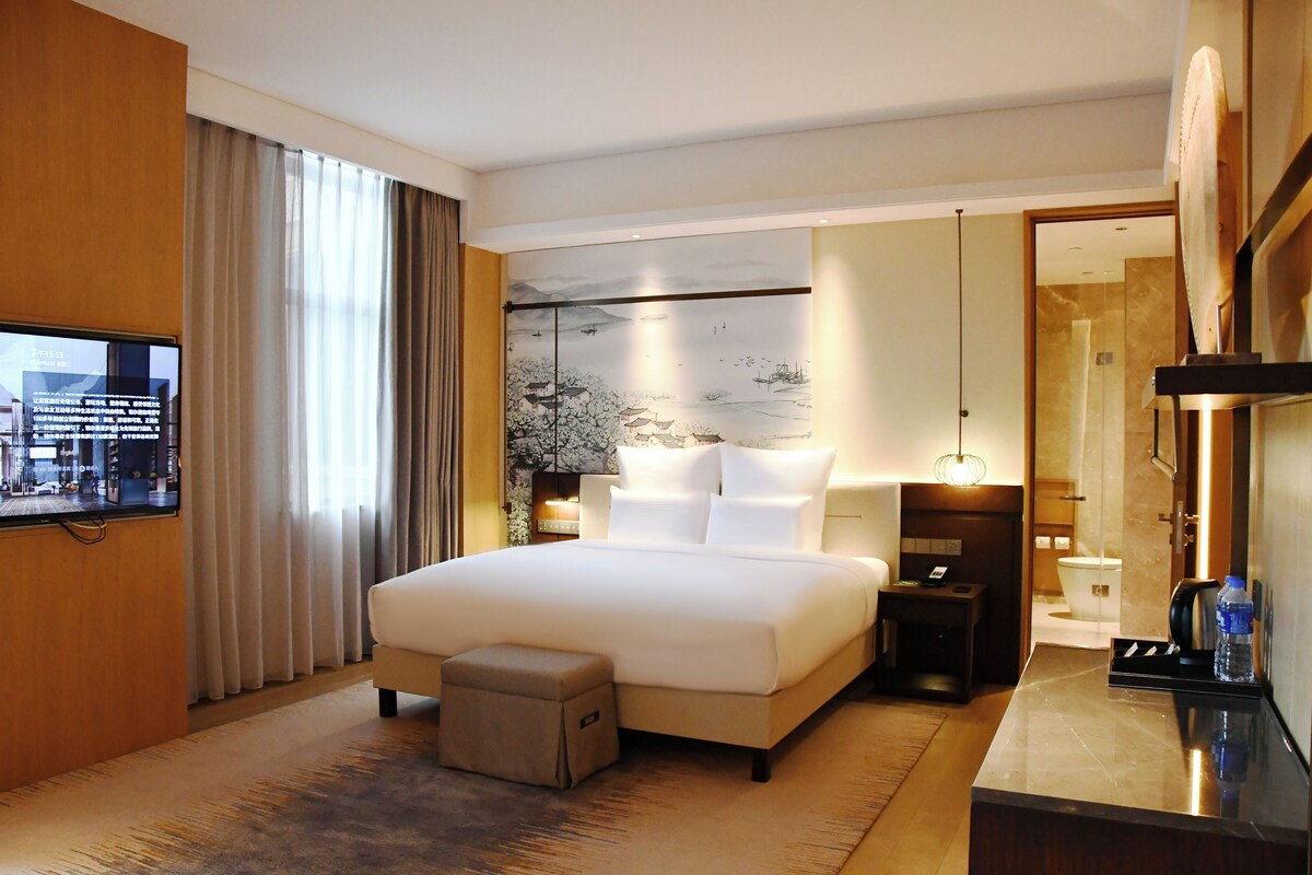 A hotel room with a large bed, a TV, a desk, and a bathroom visible through an open door.