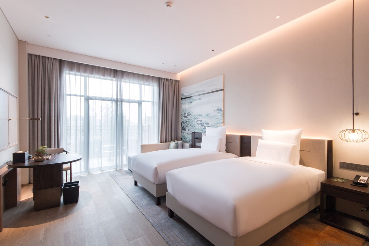 A modern hotel room with two beds, a desk, and a large window with curtains.