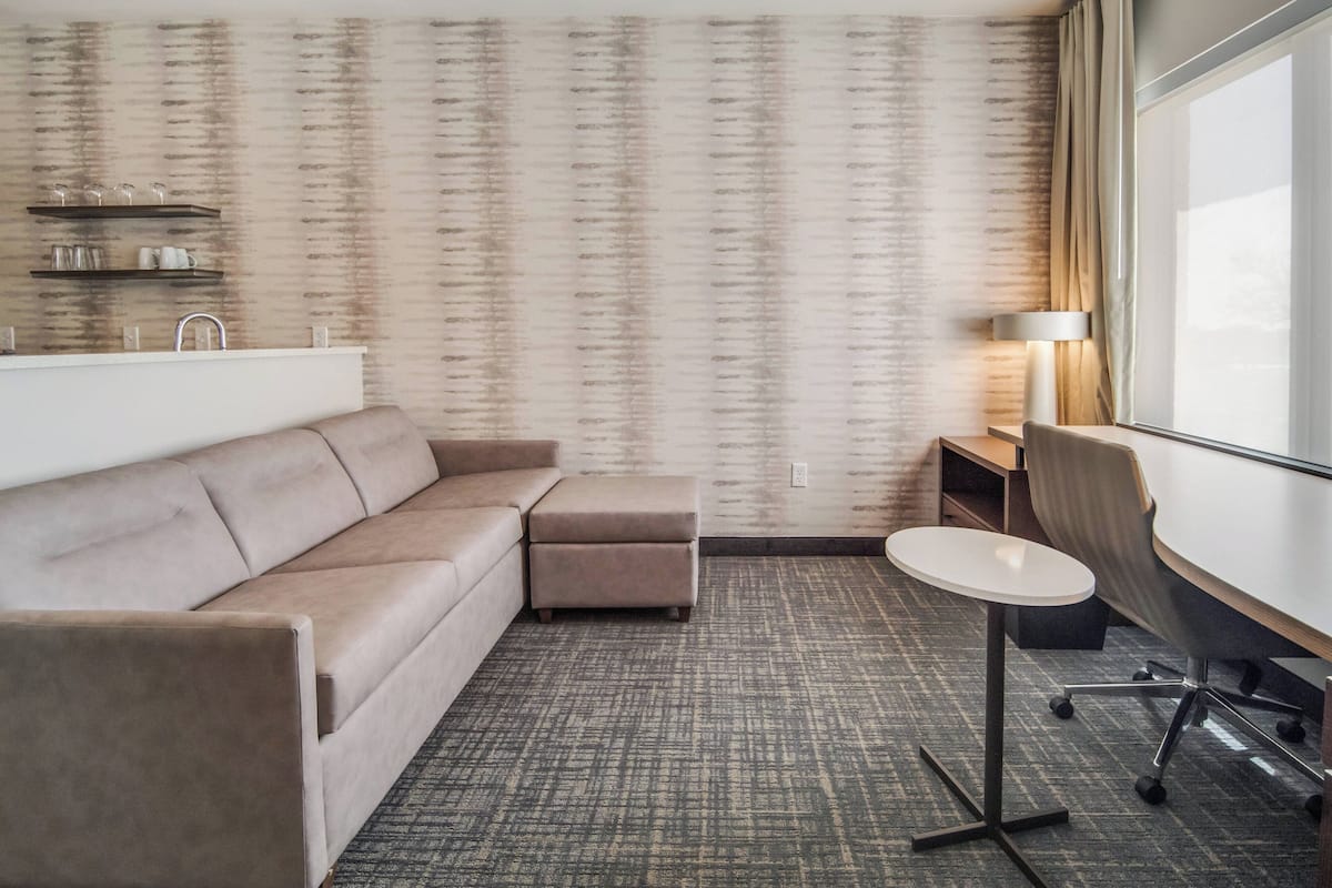 Una habitación de hotel moderna con un sofá color beige, un pequeño banquillo, una mesa redonda y un escritorio con una silla. Hay una ventana con cortinas, una lámpara y estantes con vasos y tazas.
