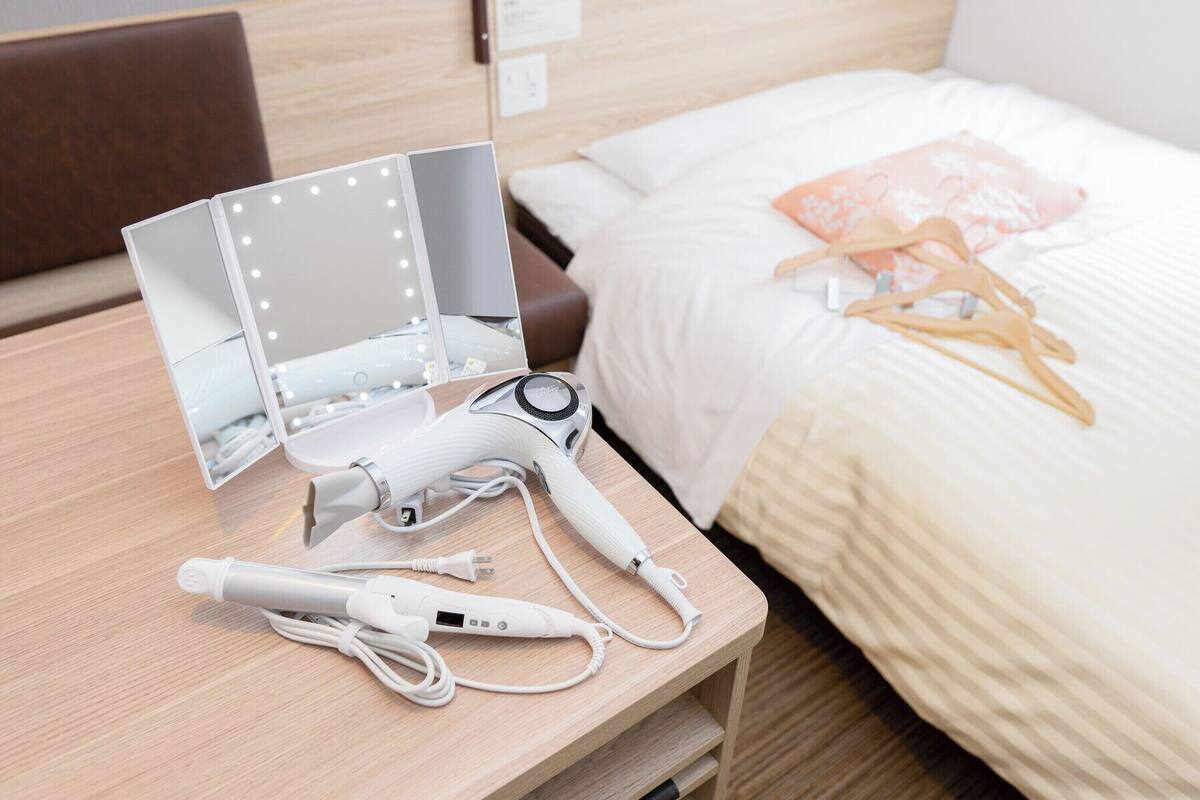 Cama bem arrumada com edredom branco e travesseiro rosa. Secador de cabelo branco e seus acessórios sobre uma mesa de cabeceira de madeira.