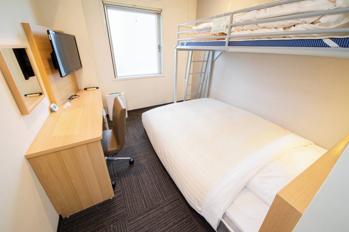 Quarto de hotel com cama de solteiro, uma escrivaninha com cadeira, televisão e janela.