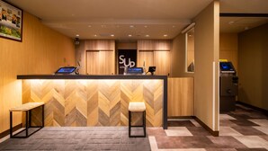 Ein Hotel-Rezeptionsbereich mit einem Holzbüro, zwei Hocker, einem digitalen Check-in-Terminal und einem Schild mit der Aufschrift 'Superhotel'.