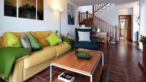 Een woonkamer met een gele bank, groene kussens, een blauwe fauteuil en een houten salontafel. Een trap met houten leuningen leidt naar een andere kamer.