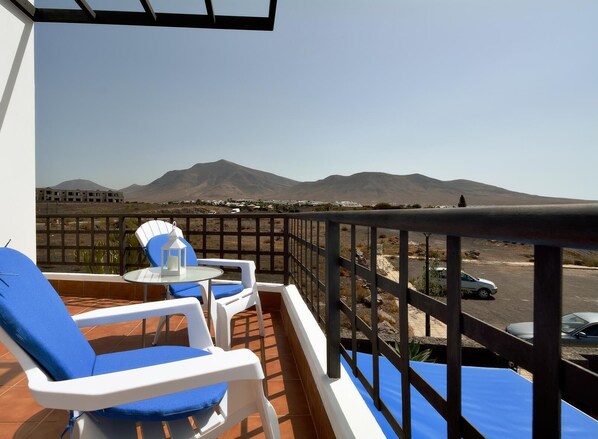 A balcony with two white chairs and a table, overlooking a mountainous landscape and a parking area. - Villa Praia Lanzarote in Playa Blanca by PVL (Playa Blanca)
