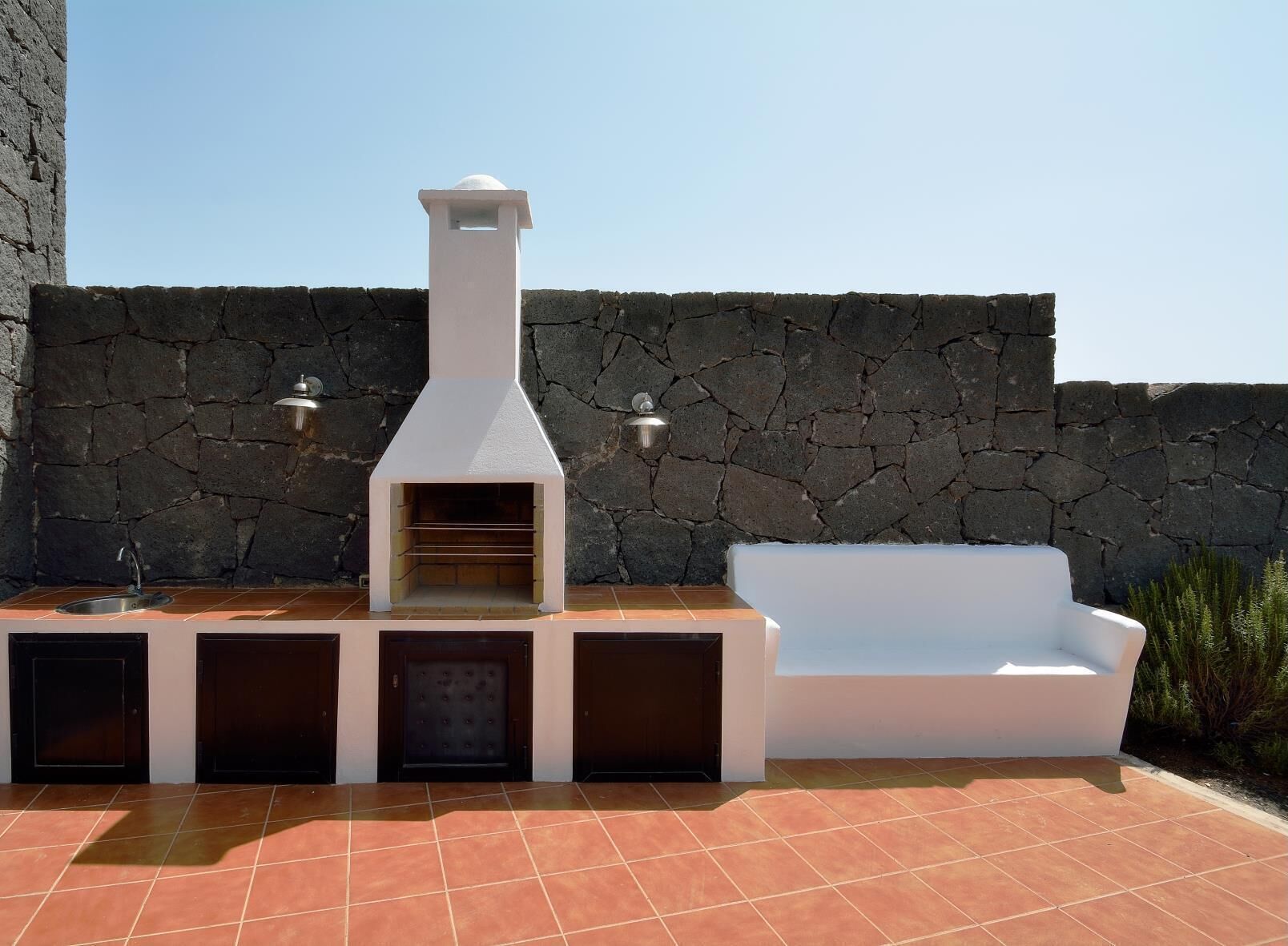 An outdoor kitchen with a stone wall backdrop, a built-in oven, and a white sofa.