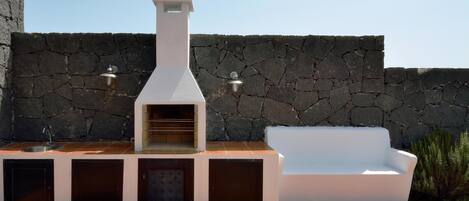 An outdoor kitchen with a stone wall backdrop, a built-in oven, and a white sofa.